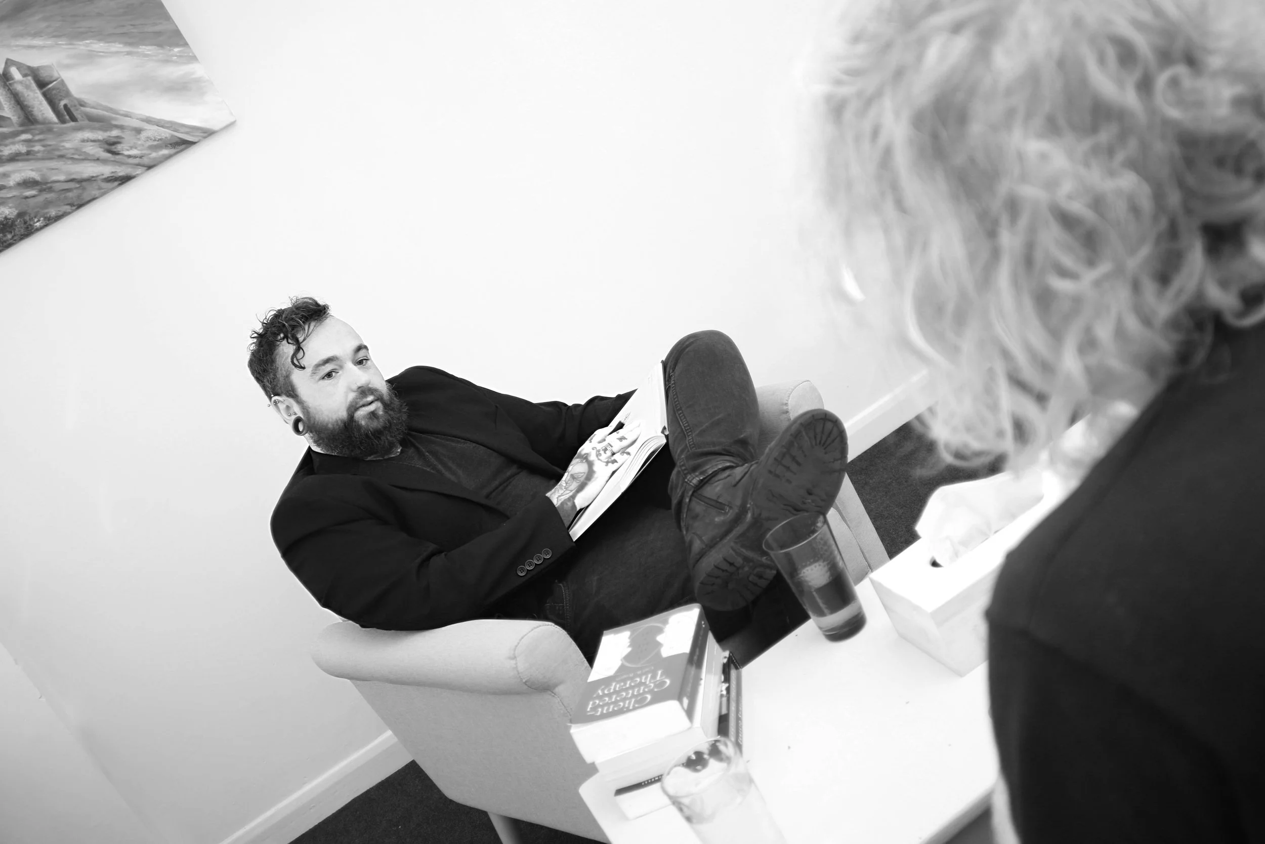 A man with a beard and tattoos, dressed in a black blazer and dark jeans, is sitting in a white armchair with his legs crossed and feet resting on a table. He is looking at an older woman with curly hair, who is seated across from him with her back to the camera. The table has a glass of water, a tissue box, a book, and a small cardboard box. There is a small part of a painting or picture on the wall above and to the left of the man.