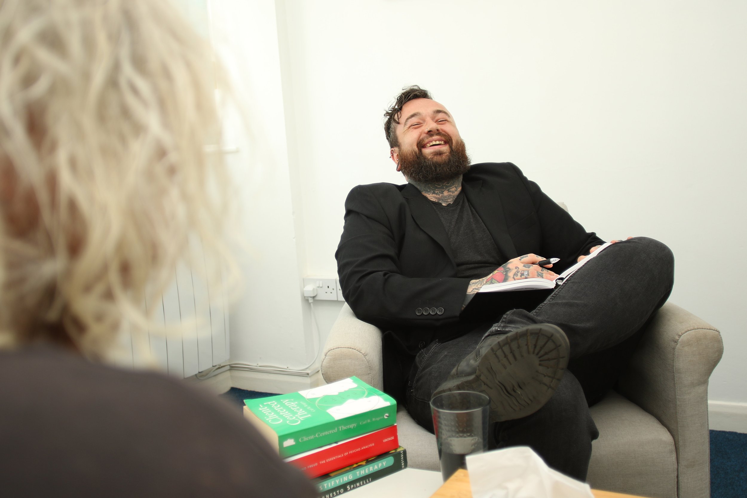 A man with tattoos and a beard, wearing a black blazer and dark pants, sitting on a small beige couch, smiling and talking during a therapy session. A woman with curly blonde hair is blurred in the foreground. Several books, a glass of water, and a tissue are visible on the table.