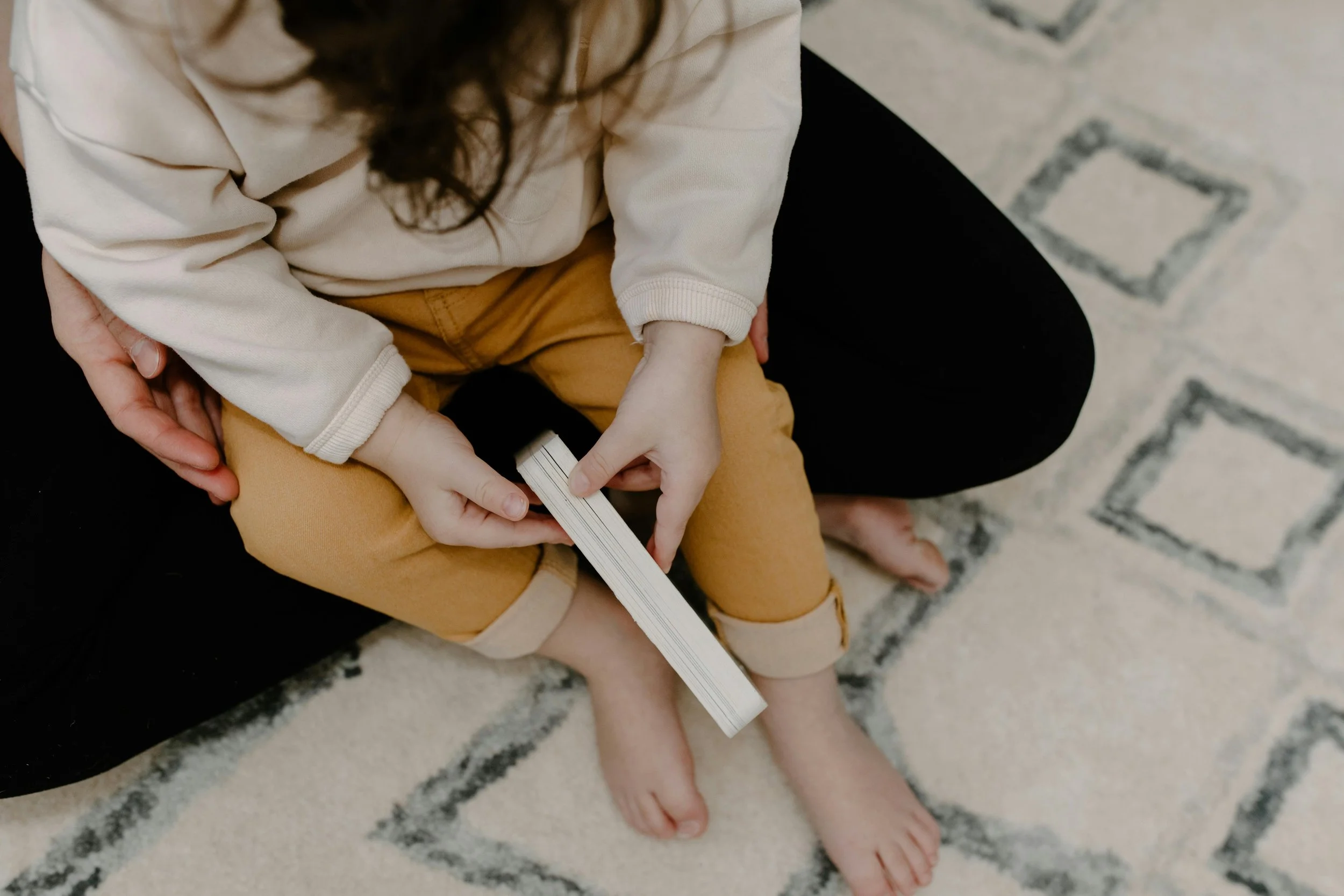 A child sitting on an adult's lap, holding a book, with a patterned rug beneath them.