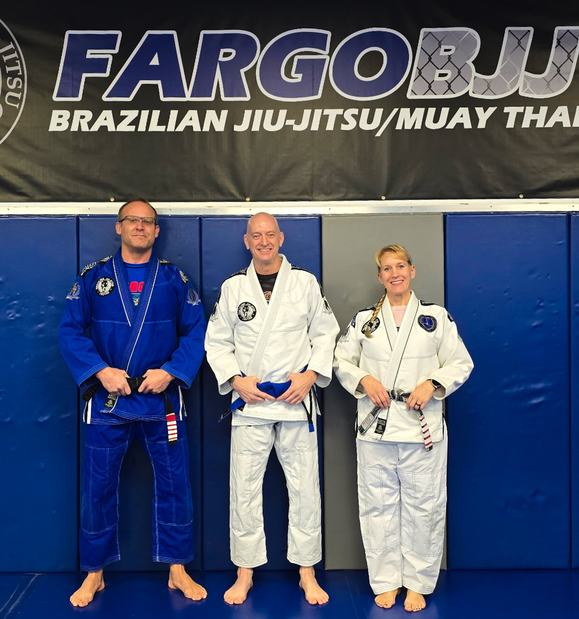 Three martial artists in gis standing on a blue mat in front of a blue padded wall, smiling, with a banner above them reading 'FARGOBJJ BRAZILIAN JIU-JITSU/MUAY THAI'