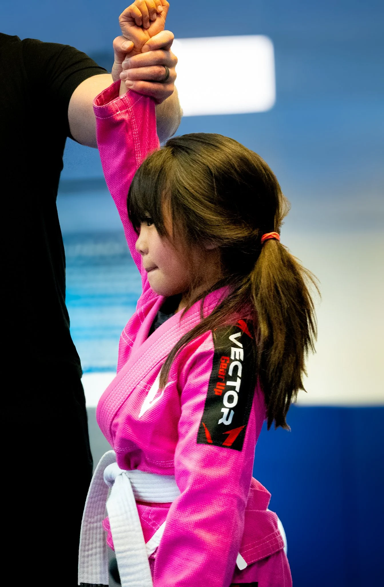 A young girl in a pink gi, with a white belt, is receiving an armbar from an instructor during a Brazilian Jiu-Jitsu class. The instructor's arm is visible, gripping her arm above the elbow.