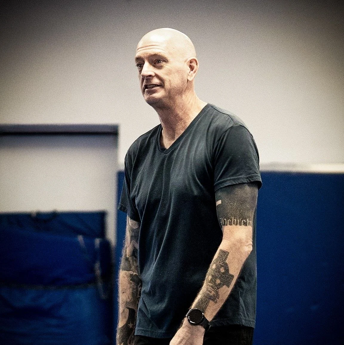 A bald man with tattoos on his arms, wearing a black t-shirt, standing indoors against a neutral background.