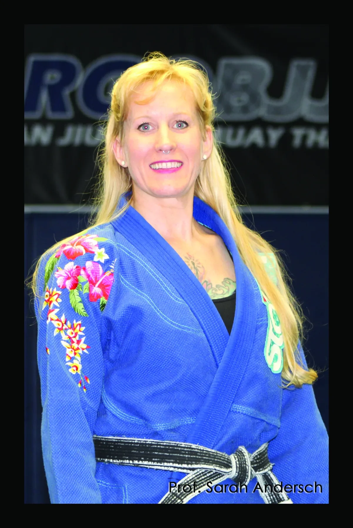 A woman with long blonde hair, wearing a blue martial arts uniform with floral embroidery on the shoulder, smiling at the camera. She has a black belt and a tattoo on her chest. The background features a black banner with blue and white text, and the text "Prof. Sarah Andersch" is at the bottom of the image.