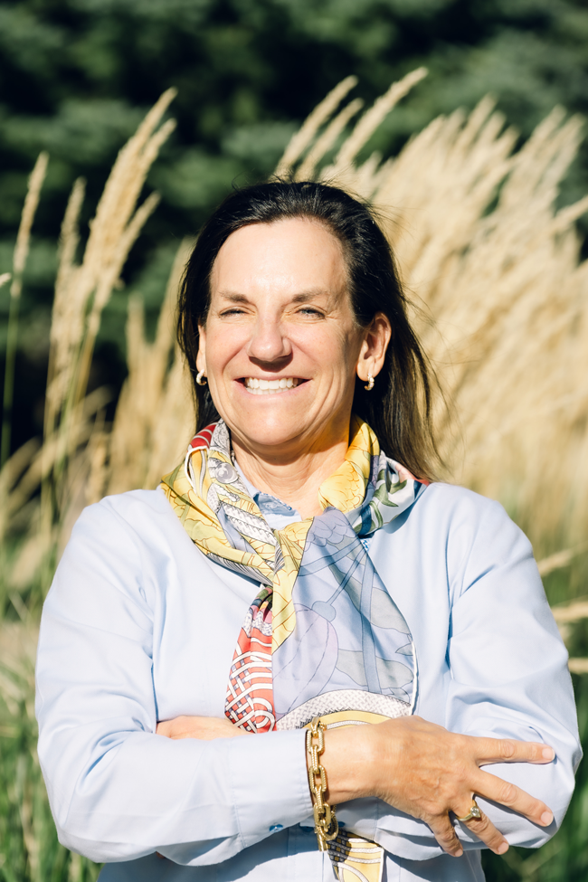 A woman smiling outdoors with tall grass and trees in the background.