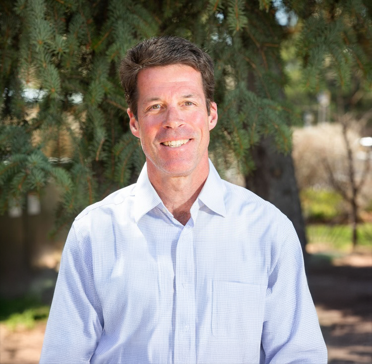 A smiling man in a white button-up shirt standing outdoors near a large tree.