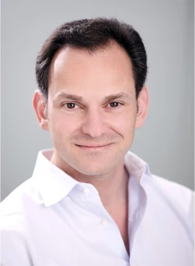 Headshot of a man with dark hair, smiling, wearing a white collared shirt against a blurred gray background.