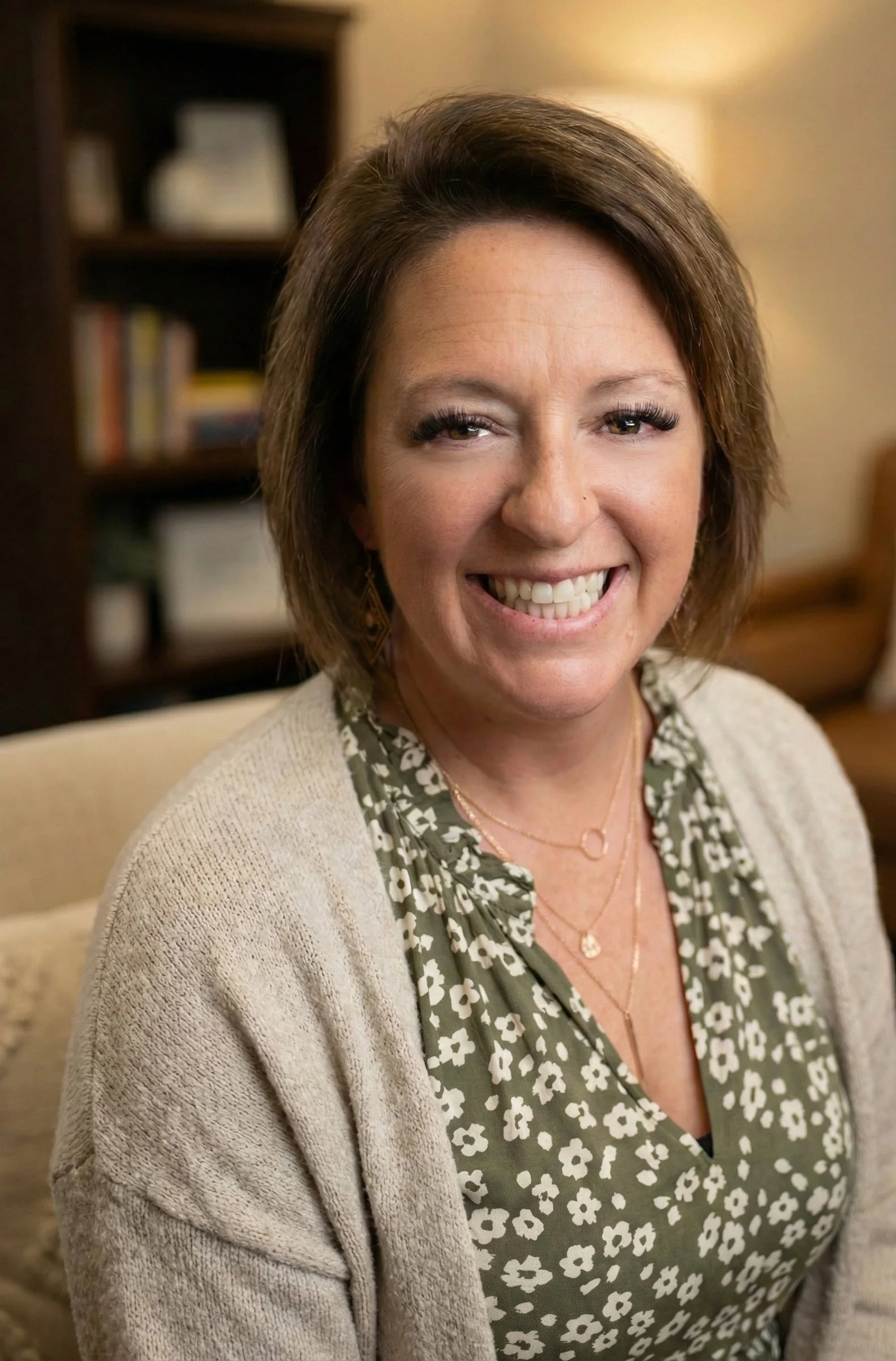 A woman with shoulder-length brown hair smiling, wearing a green floral blouse and a beige cardigan, seated in a cozy room with a bookshelf in the background.