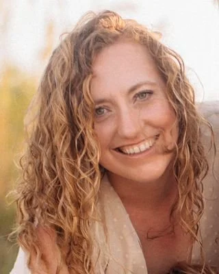 A woman with curly blonde hair smiling outdoors with a blurred background.