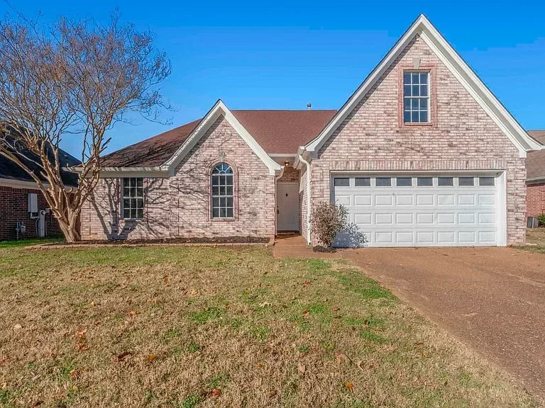 Front view of a two-story house with a brick exterior, a brown shingle roof, front yard with grass and a leafless tree, driveway leading to a two-car garage, and a small front pathway.