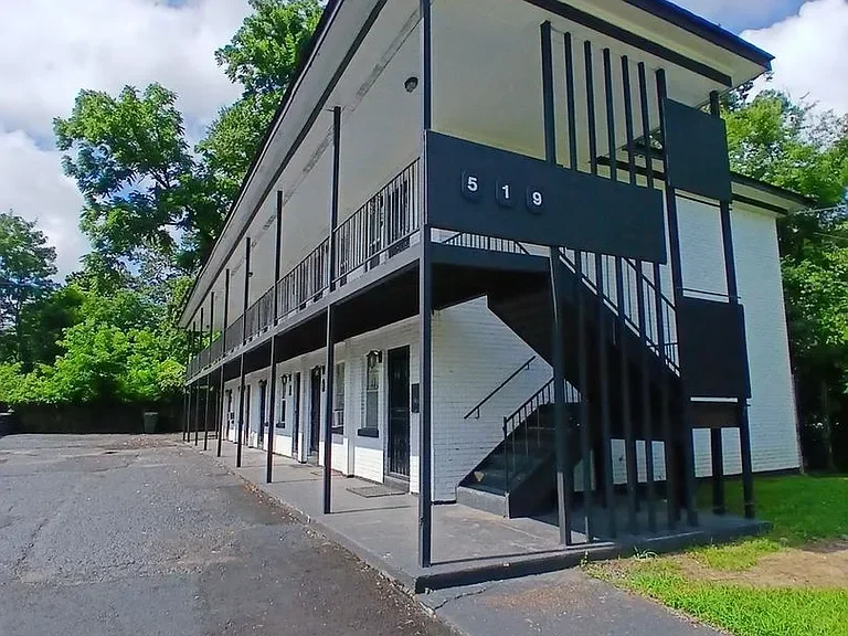 A two-story apartment building with white exterior walls and black trim, featuring a black metal staircase on the side and a large black sign with the number 519. The building has exterior hallways and multiple doors, with trees and greenery in the background.