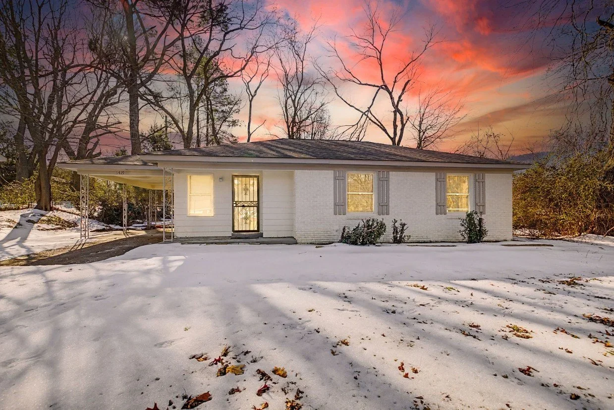 A single-story white house with gray shutters, lit windows, and a carport on the left, surrounded by snow-covered ground and leafless trees at sunset with colorful sky.