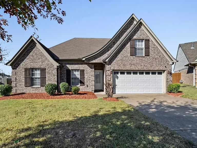 Front view of a brick house with a garage, two windows with shutters, and a small front yard with bushes and grass.