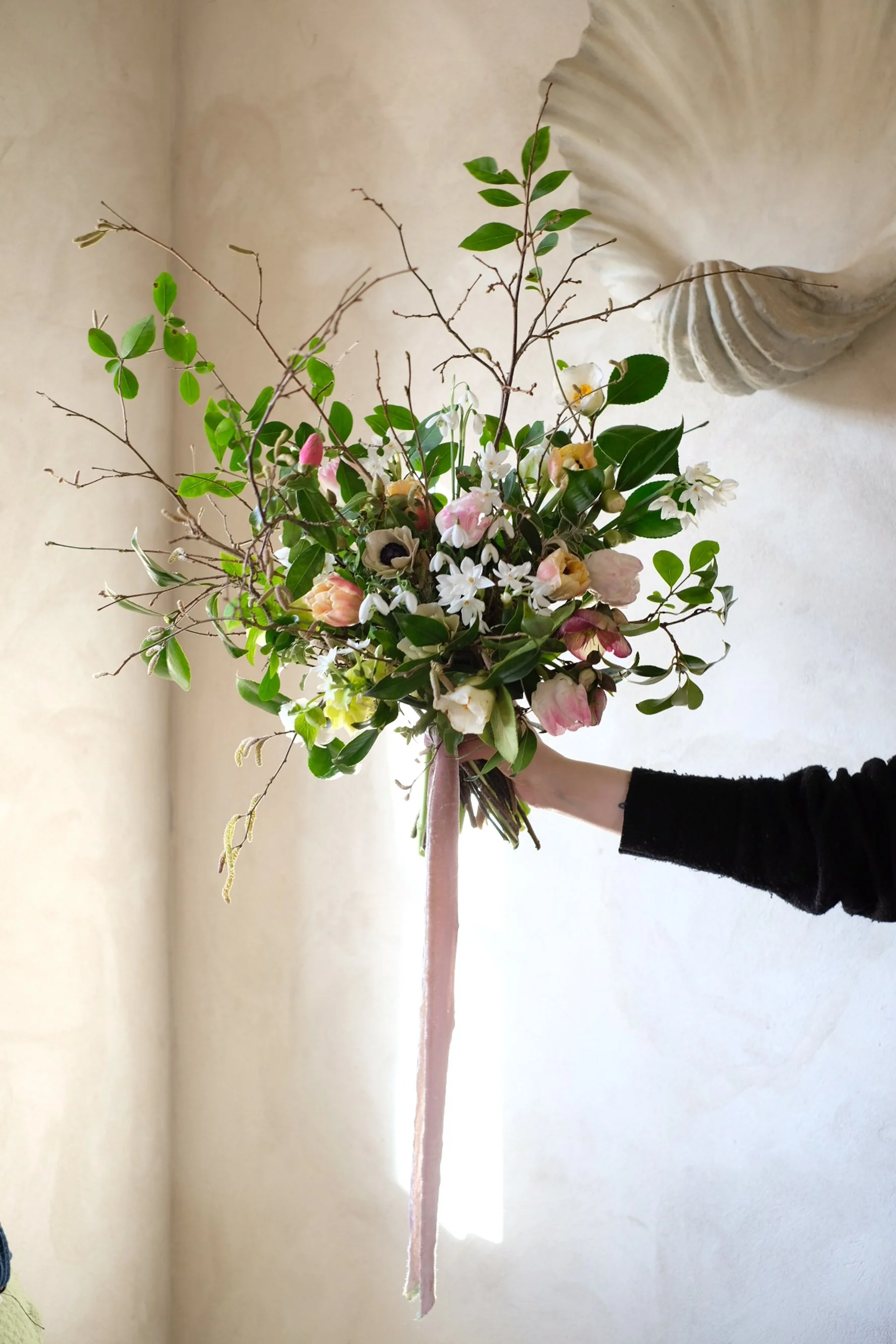 A person holding a large bouquet of mixed flowers with green leaves, pink and white blooms, and some twisted branches, against a light beige indoor wall.