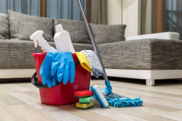 Cleaning supplies including a red bucket with a spray bottle, gloves, a mop, and cleaning cloths on hardwood floor in front of a sofa.