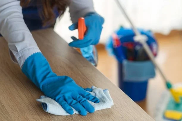 Someone cleaning a wooden surface with a cloth and spray bottle, wearing blue gloves.