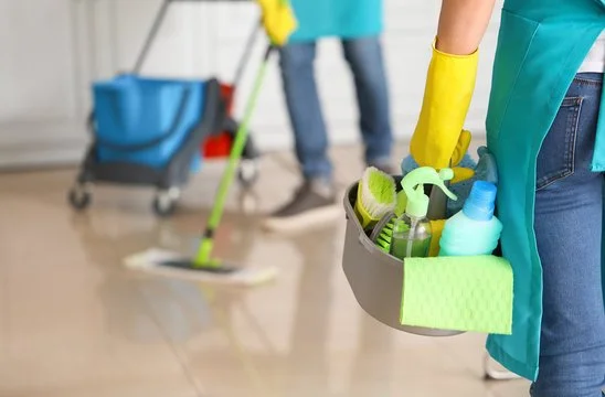 Person holding a cleaning caddy with spray bottles, sponges, and cleaning supplies, with another person cleaning the floor in the background.