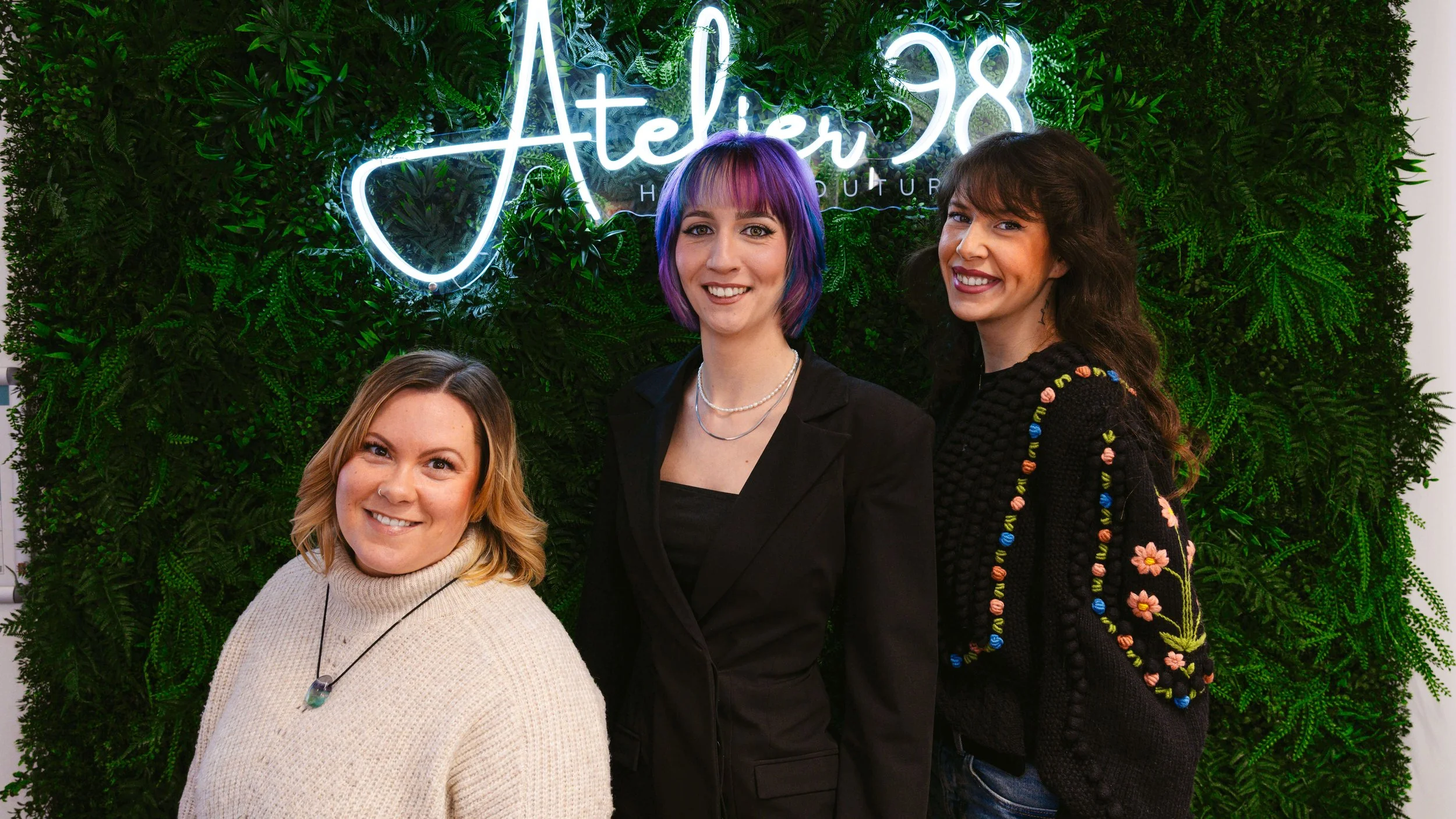 Drei Frauen stehen vor einer grünen Wand mit einem Neon-Schild, das sagt "Atelier 98'.