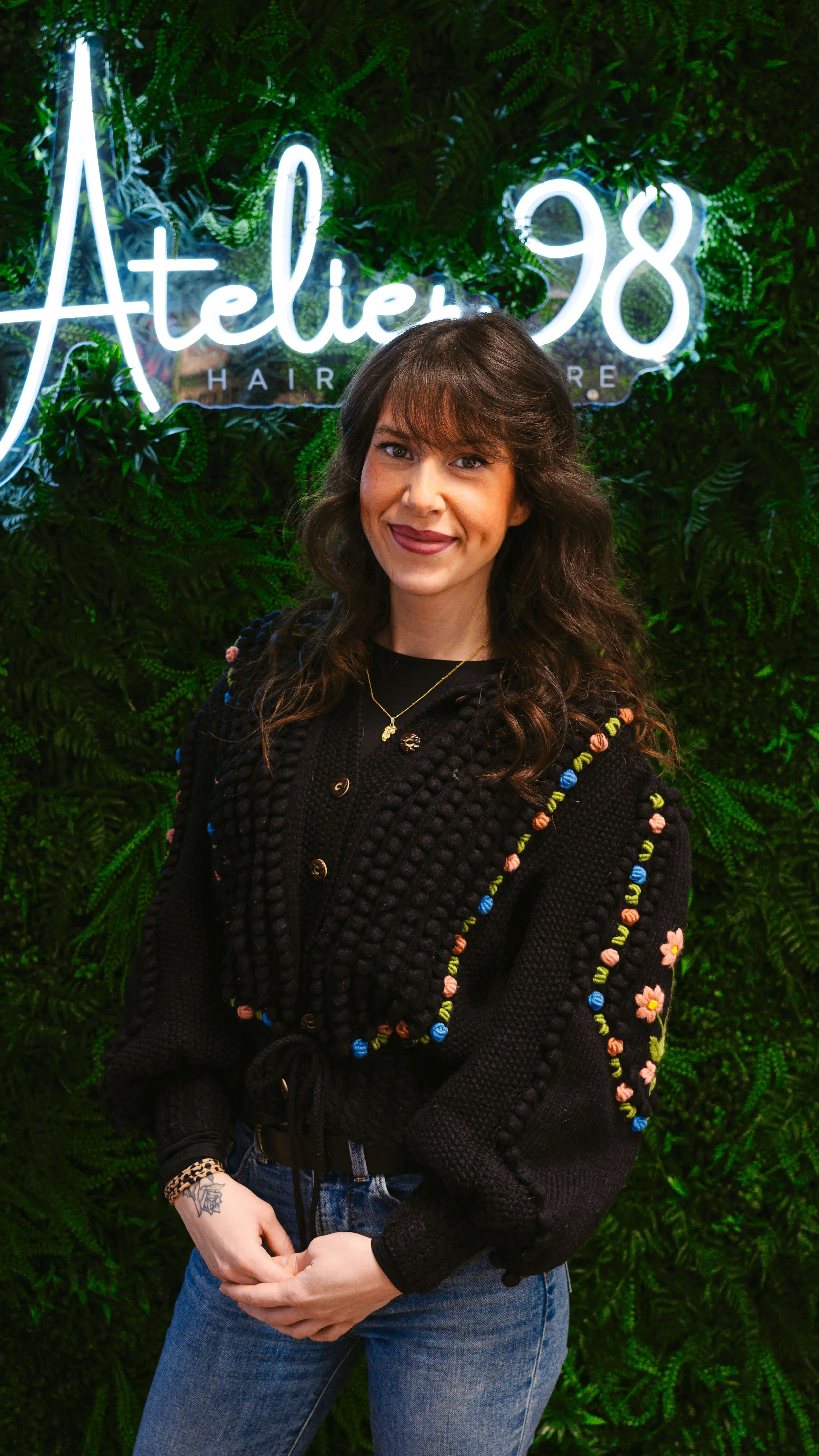 Junge Frau mit dunklen, gewellten Haaren und dunkler Kleidung, die vor einem grünen Pflanzenhintergrund steht und lächelnd in die Kamera schaut, neben einem Neon-Schild, das 'Atelier 98 Hair' zeigt.