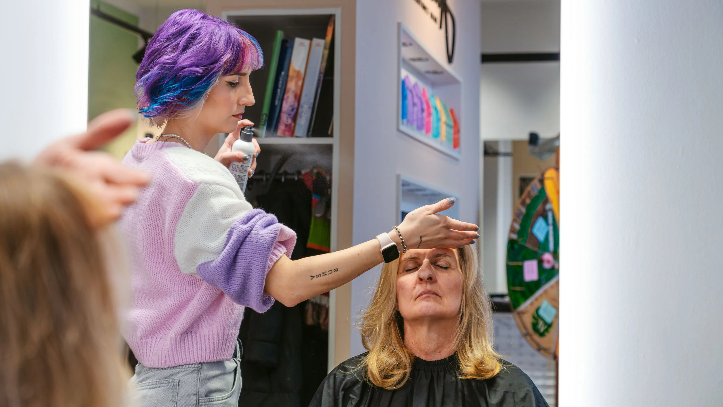 Eine Friseurin sprüht Haarspray auf einen Kunden, der mit geschlossenen Augen sitzt, während eine weitere Person im Vordergrund zuschaut.