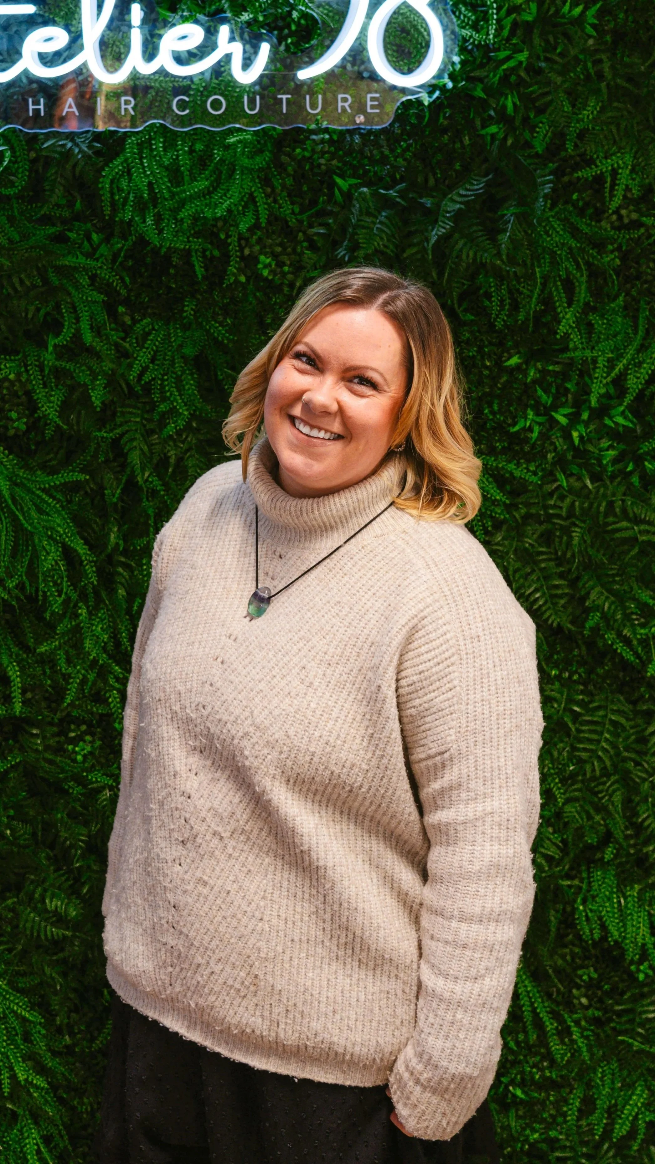Eine lächelnde Frau mit blonden, schulterlangen Haaren trägt einen beigen Rollkragenpullover und einen Halsreif mit Anhänger, im Hintergrund eine grüne Wand mit Pflanzen und einem leuchtenden Neon-Schild.