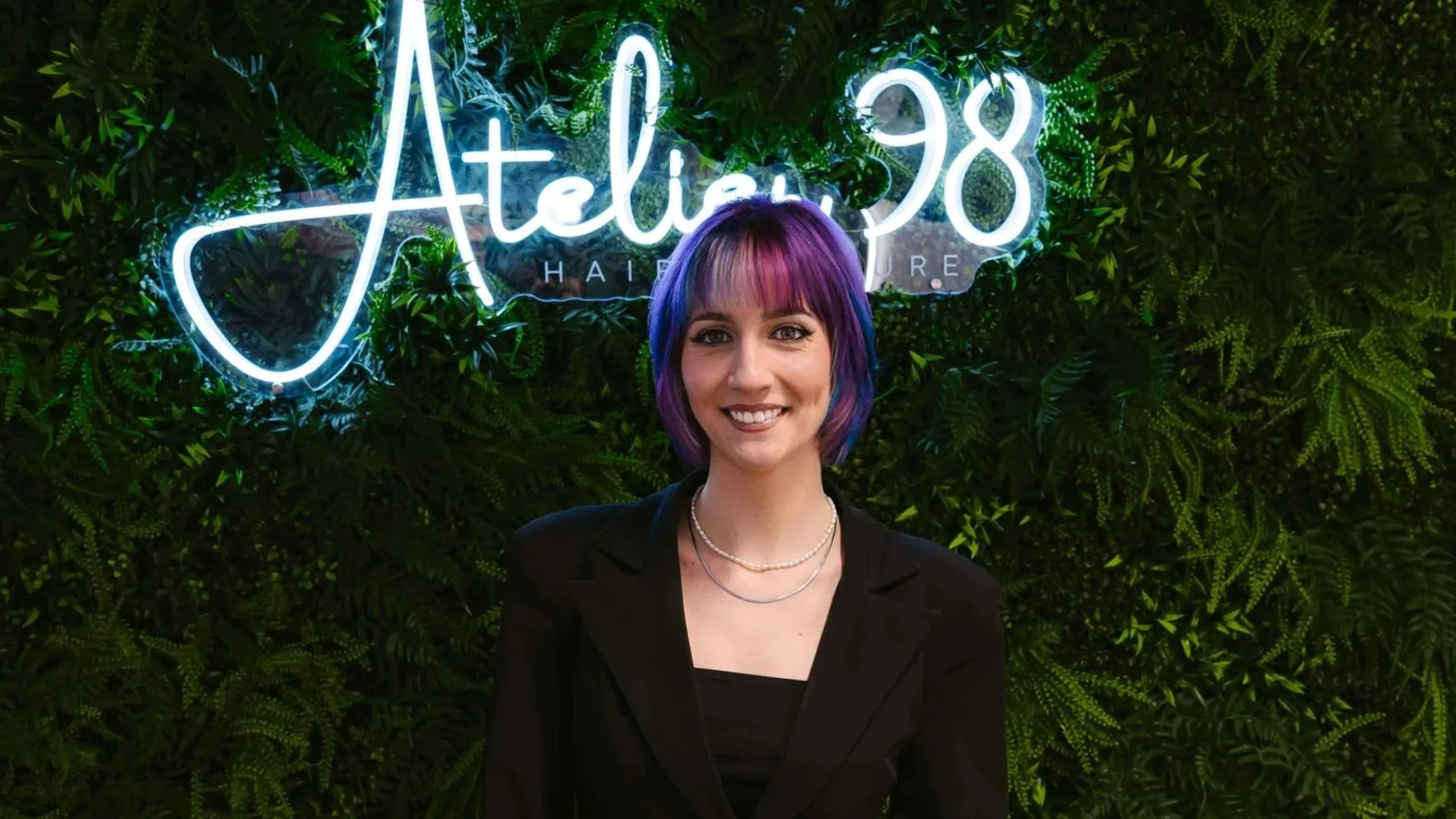 Eine junge Frau mit bunter, mehrfarbiger Frisur (Lila, Blau, Pink) vor einer grünen Wand mit Neon-Schriftzug "Jatelin 98" und Blättern.