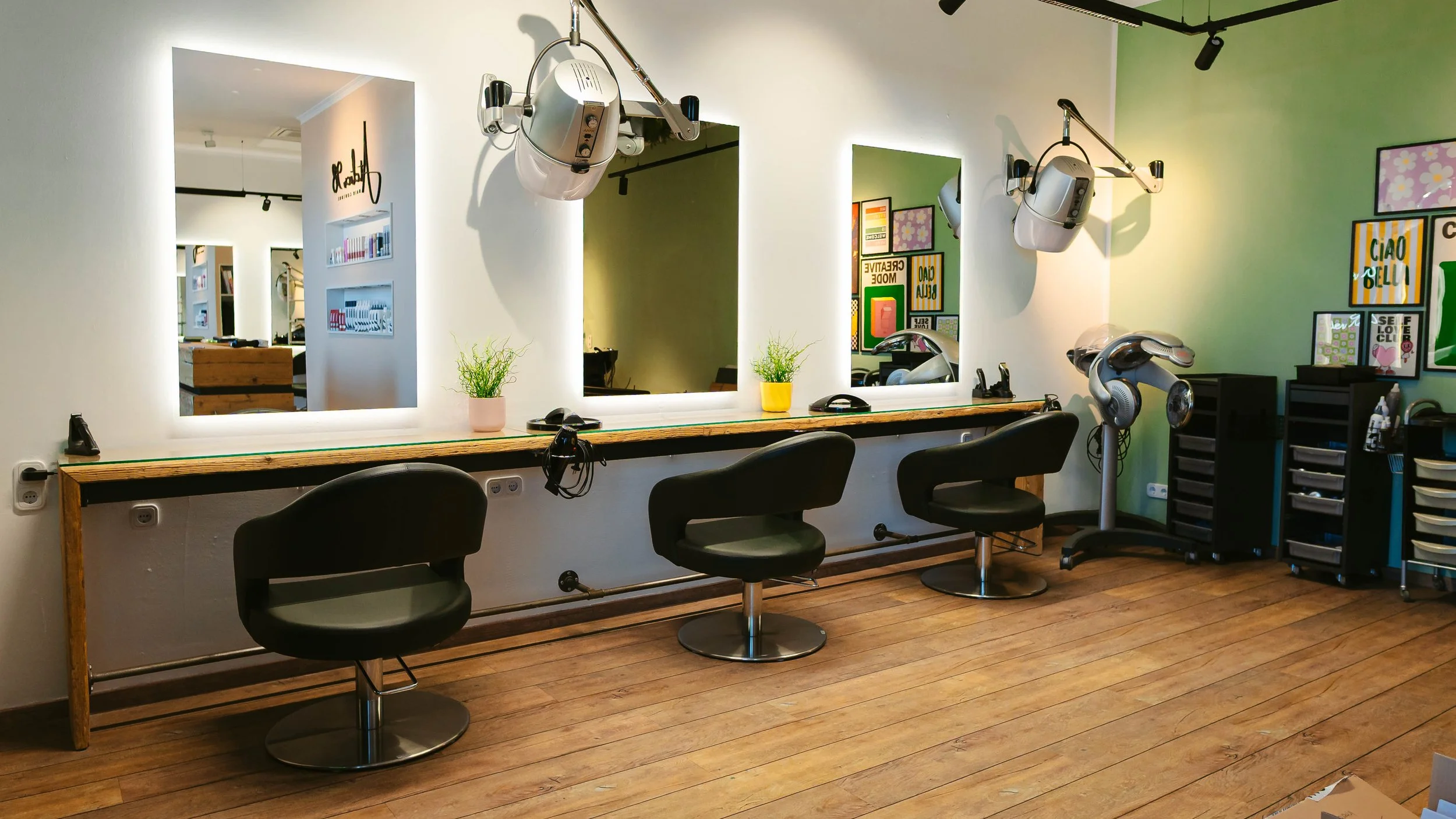 Hintere Perspektive eines Friseursalons mit drei schwarzen Friseurstühlen vor einem langen Spiegel, auf dem zwei Pflanzen in Töpfen stehen. Im Hintergrund sind Bilder und Poster auf der grünen Wand und Haarstylinggeräte wie Haartrockner und Lockenstab. Der Boden ist aus Holzplanken.