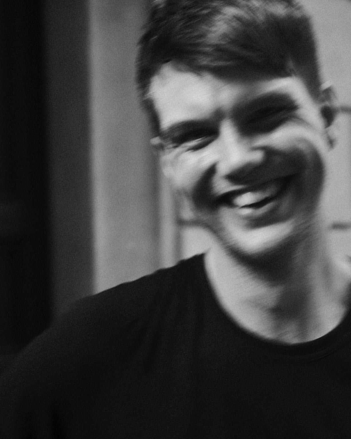 A black and white photo of a smiling young man with short hair, wearing a dark shirt.