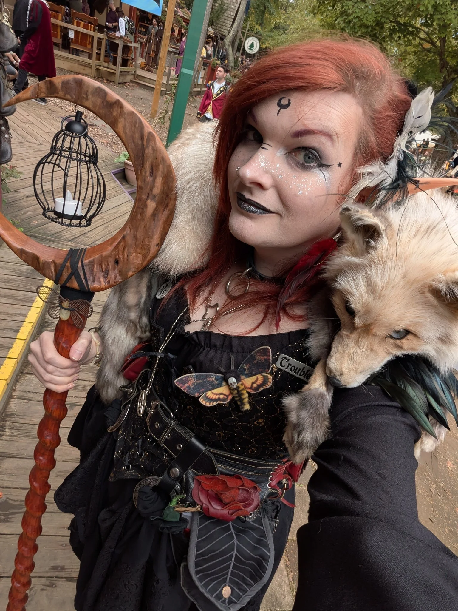 A woman with red hair dressed in fantasy-style costume holding a staff with a cage at the top. She has face makeup with a moon symbol on her forehead, glitter, and dark lipstick. She is carrying a small animal, possibly a fox, on her shoulder. She is outdoors at a themed event or festival with wooden walkways and other people in the background.