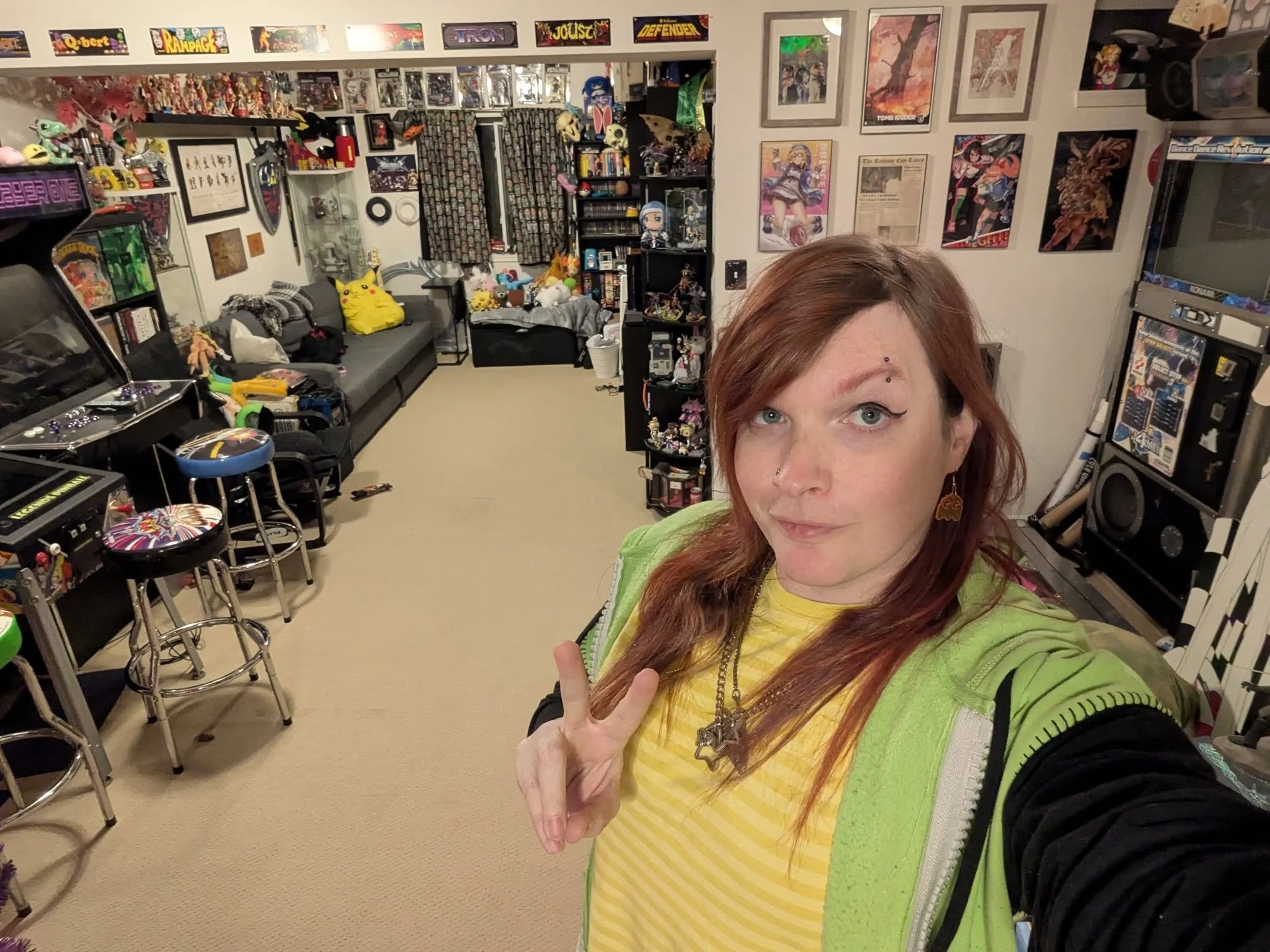 A woman with reddish hair, wearing a yellow shirt and green jacket, taking a selfie in a cluttered living room decorated with framed posters and anime figures.