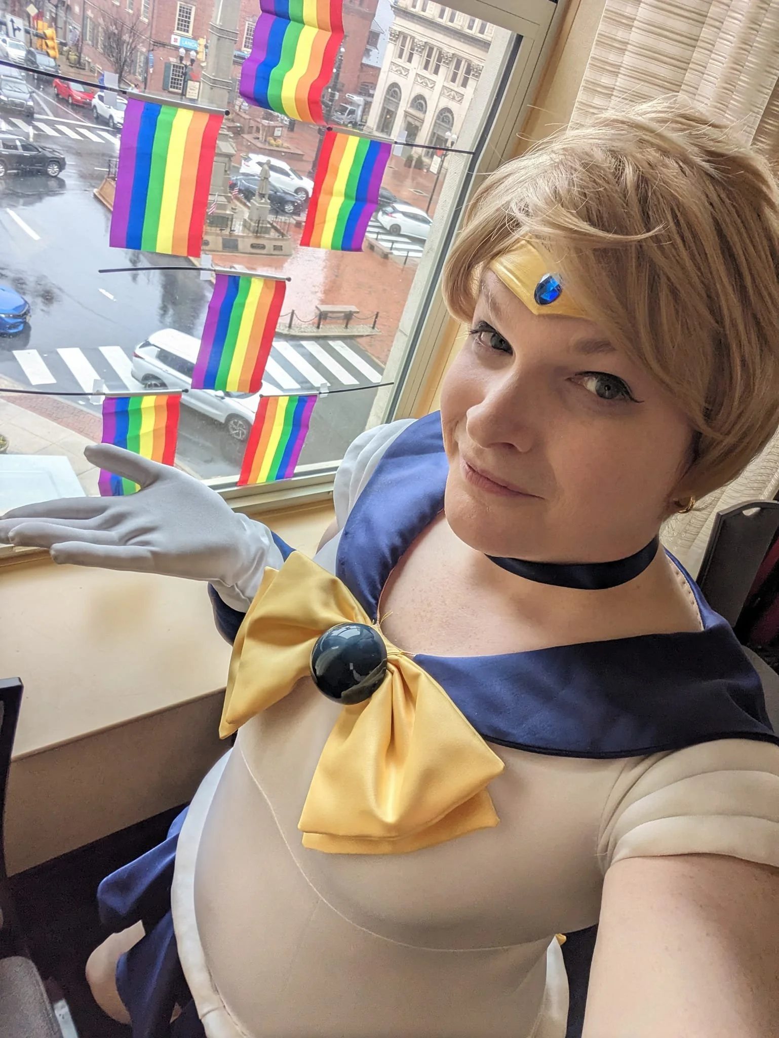 Person dressed as Sailor Moon posing indoors near a window with rainbow pride flags outside.