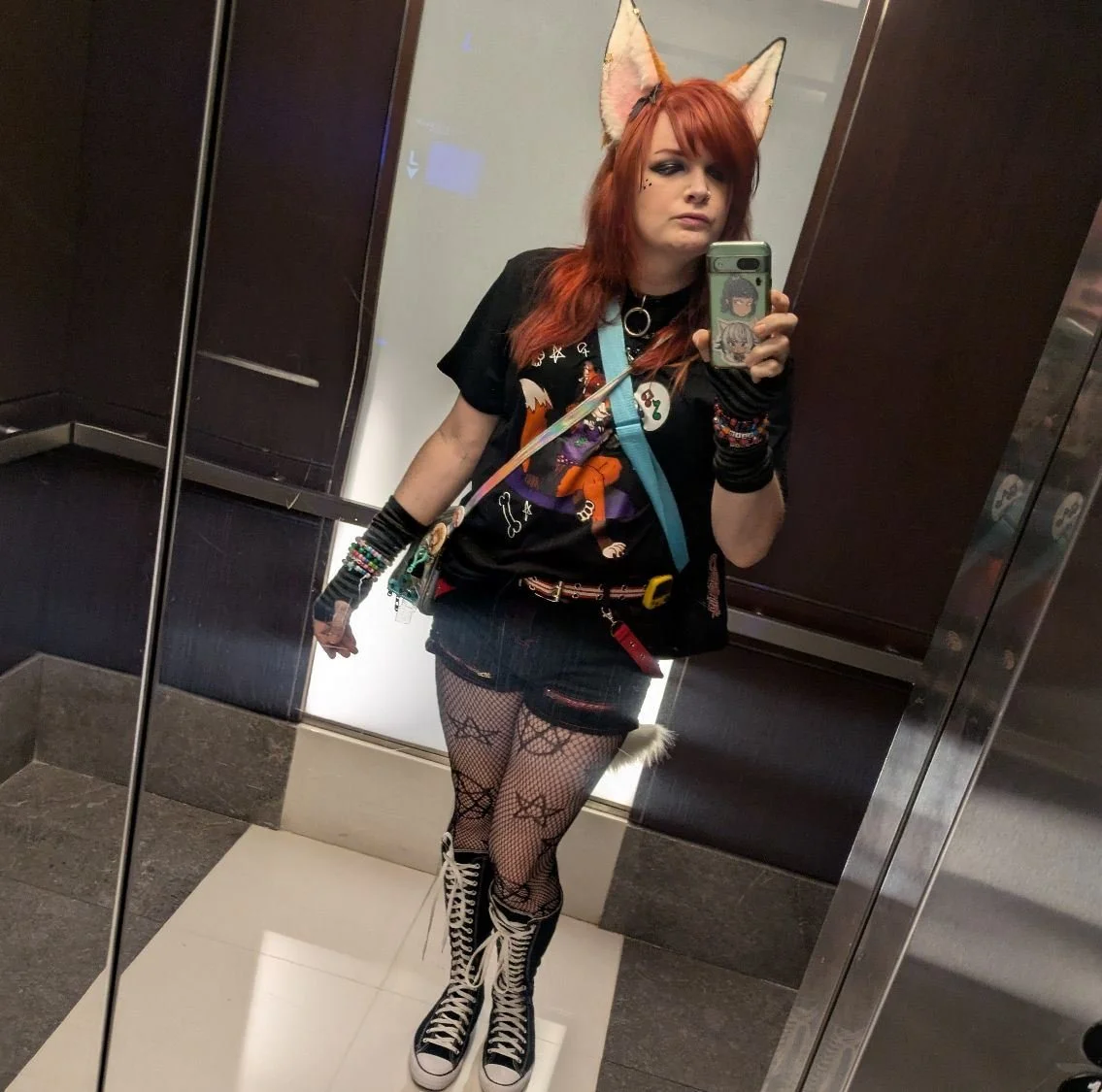 Woman with red hair, fox ears headband, black graphic t-shirt, fishnet stockings with cat paw patterns, and high-top sneakers taking a selfie in an elevator mirror.