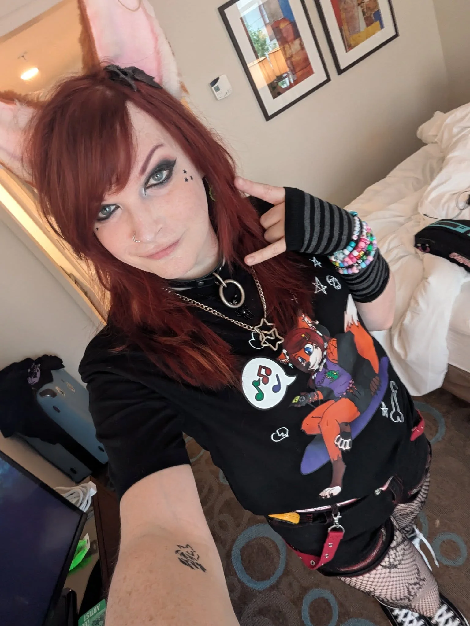 Young woman with red hair wearing fox ears headband, black graphic t-shirt, fishnet stockings, and colorful bracelets, taking a selfie in a hotel room with bed, artwork, and a dresser visible in the background.