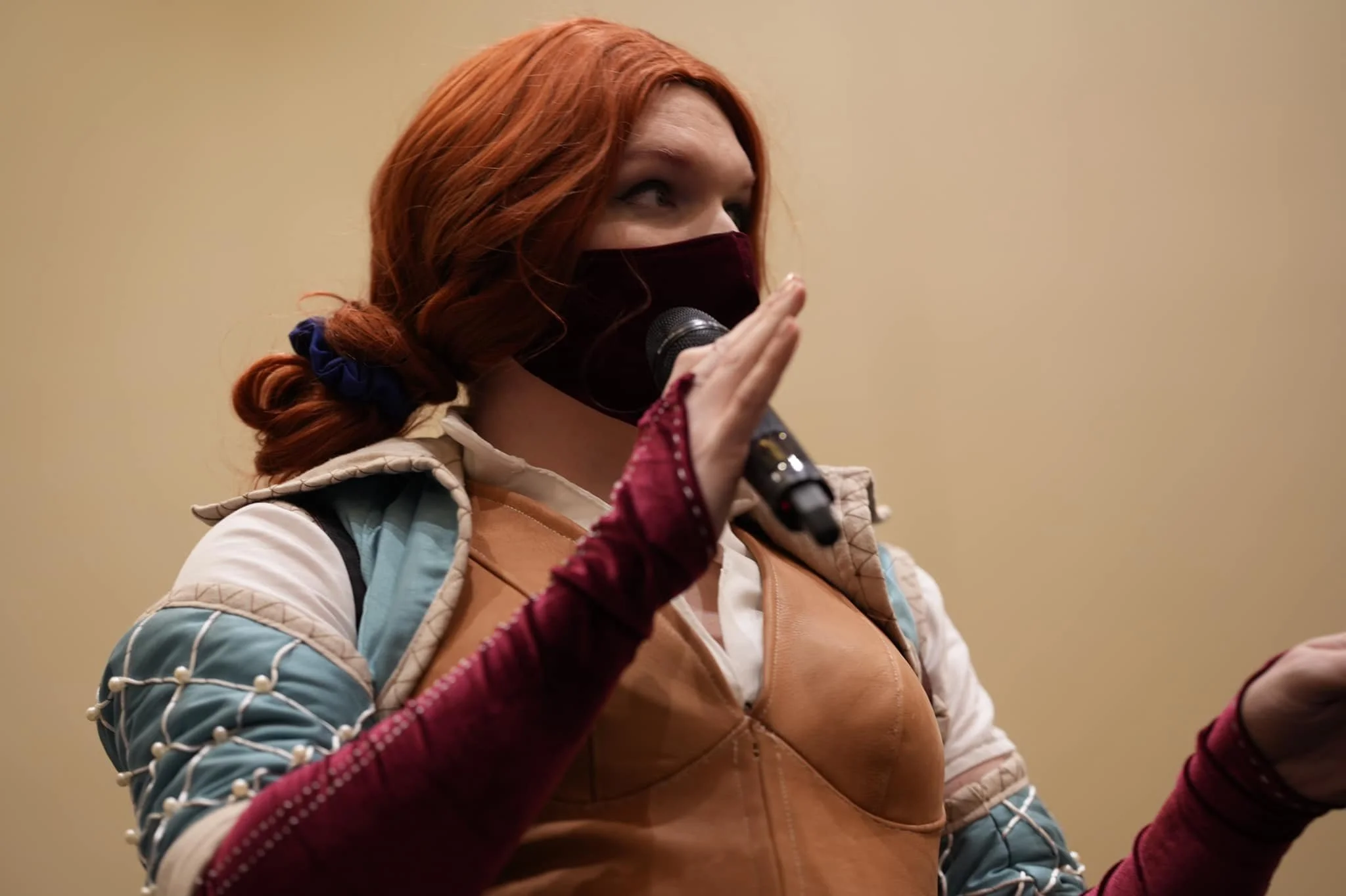 A woman with red hair, wearing a face mask, holding a microphone, and gesturing with her hand.