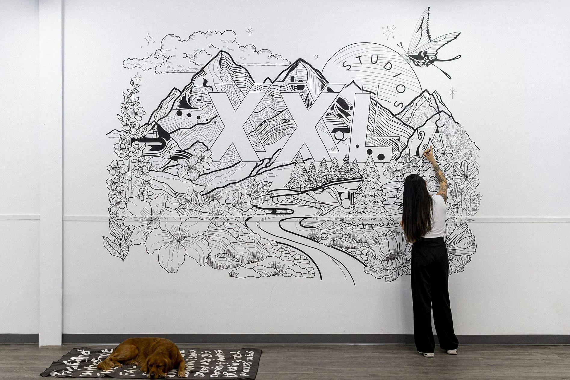 A woman with long dark hair drawing on a large mural of a mountain landscape with flowers, trees, and a river. A dog is sleeping on a mat on the floor nearby.