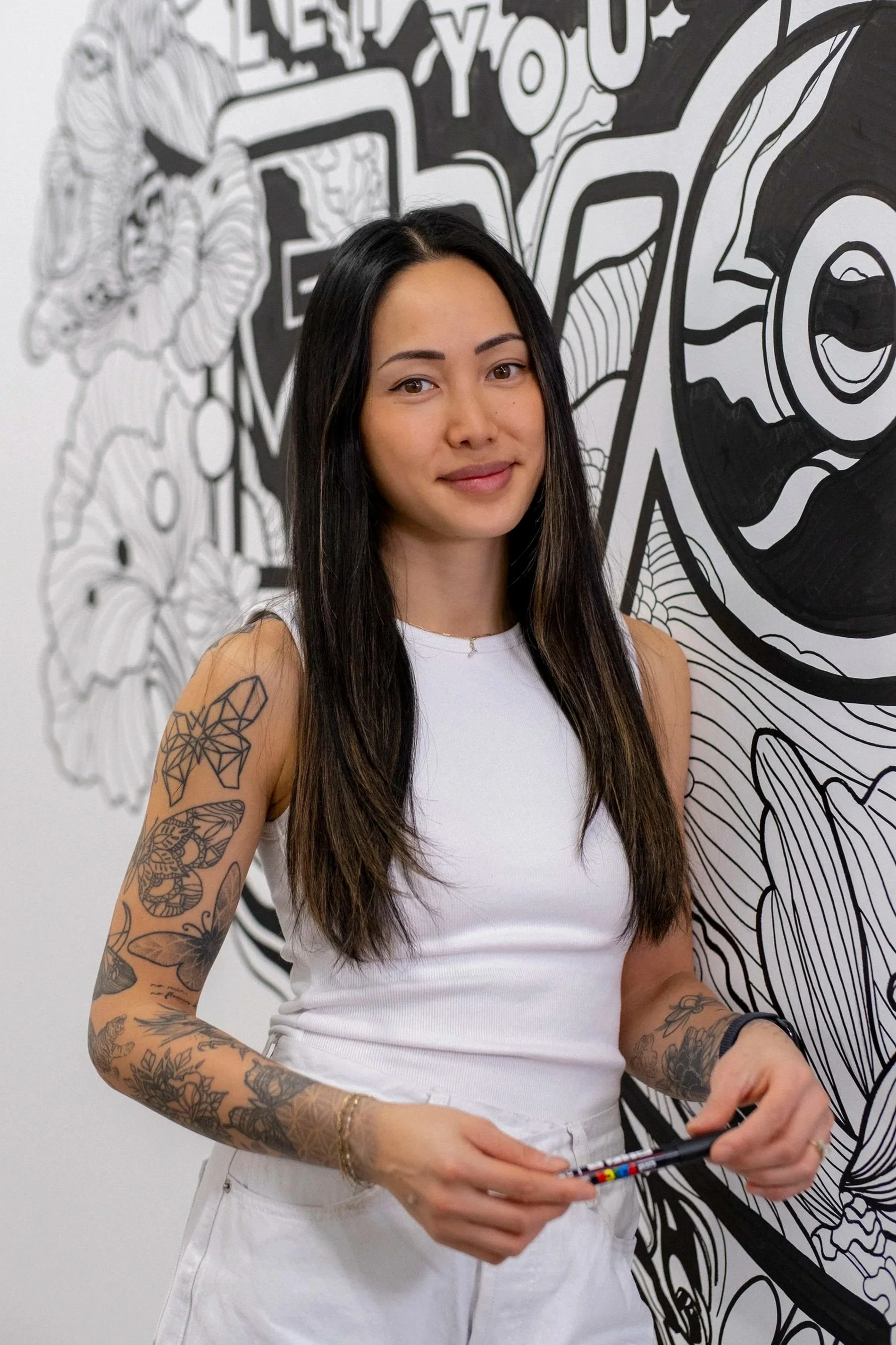 A young woman with long dark hair and tattoos on her arms standing in front of a black and white floral and abstract mural, holding a marker.