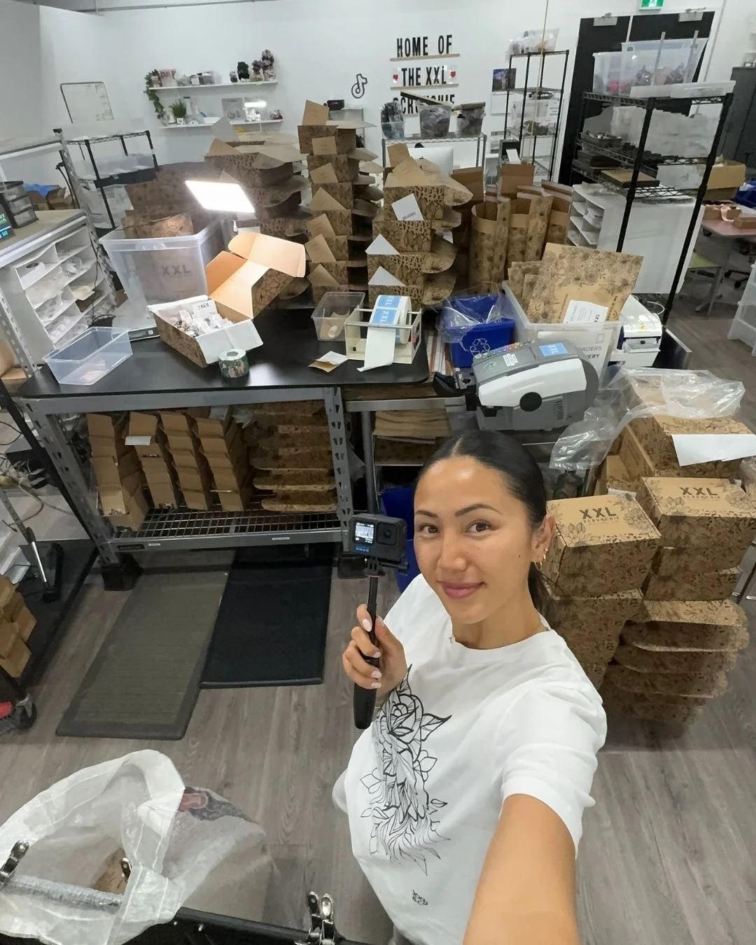 A woman taking a selfie in a workspace with many cardboard boxes and packaging materials on tables and shelves surrounding her.