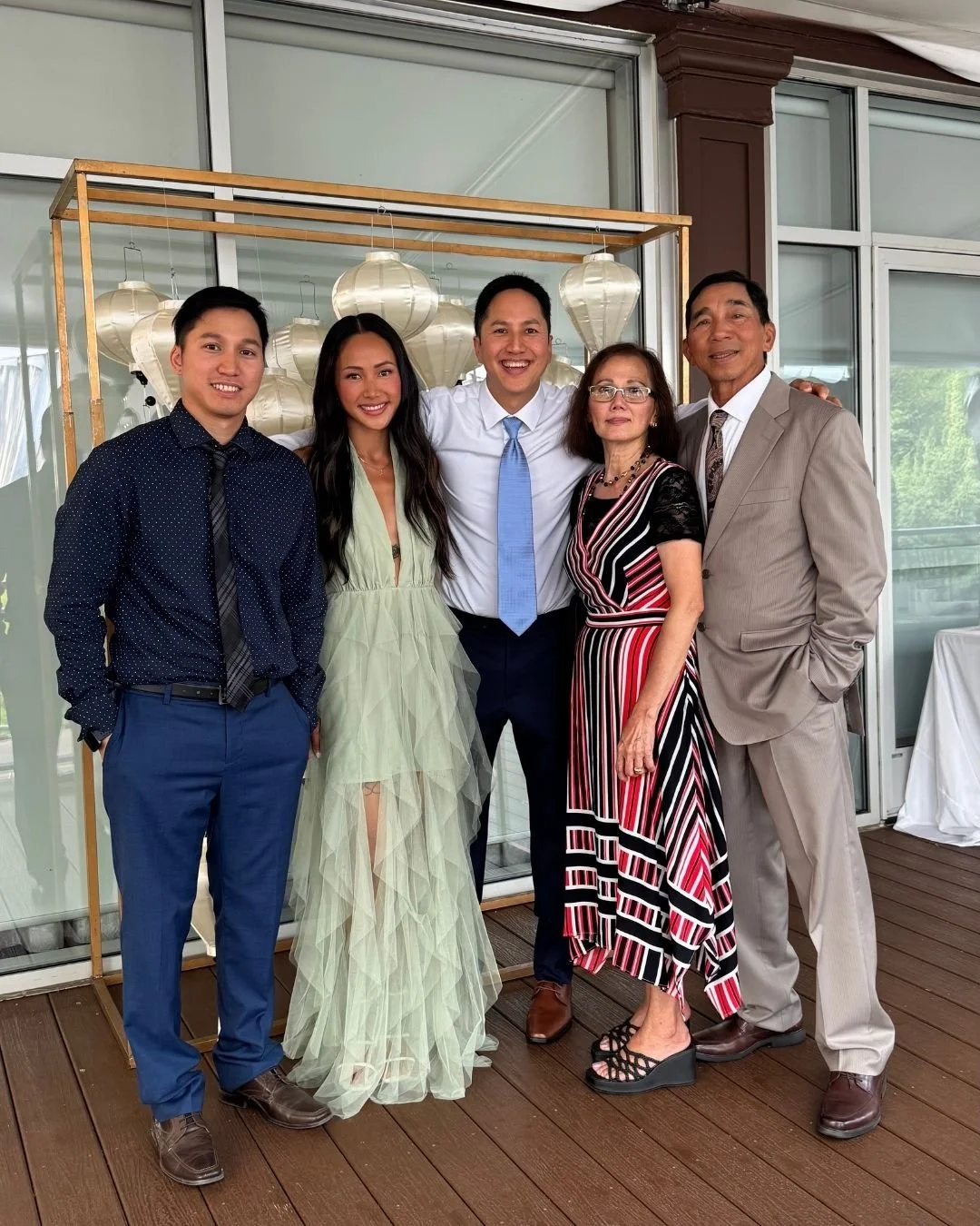 Group of five people, three men and two women, dressed in formal attire, standing on a wooden deck in front of glass doors, smiling for a photo. There are white lanterns and a wooden frame in the background.
