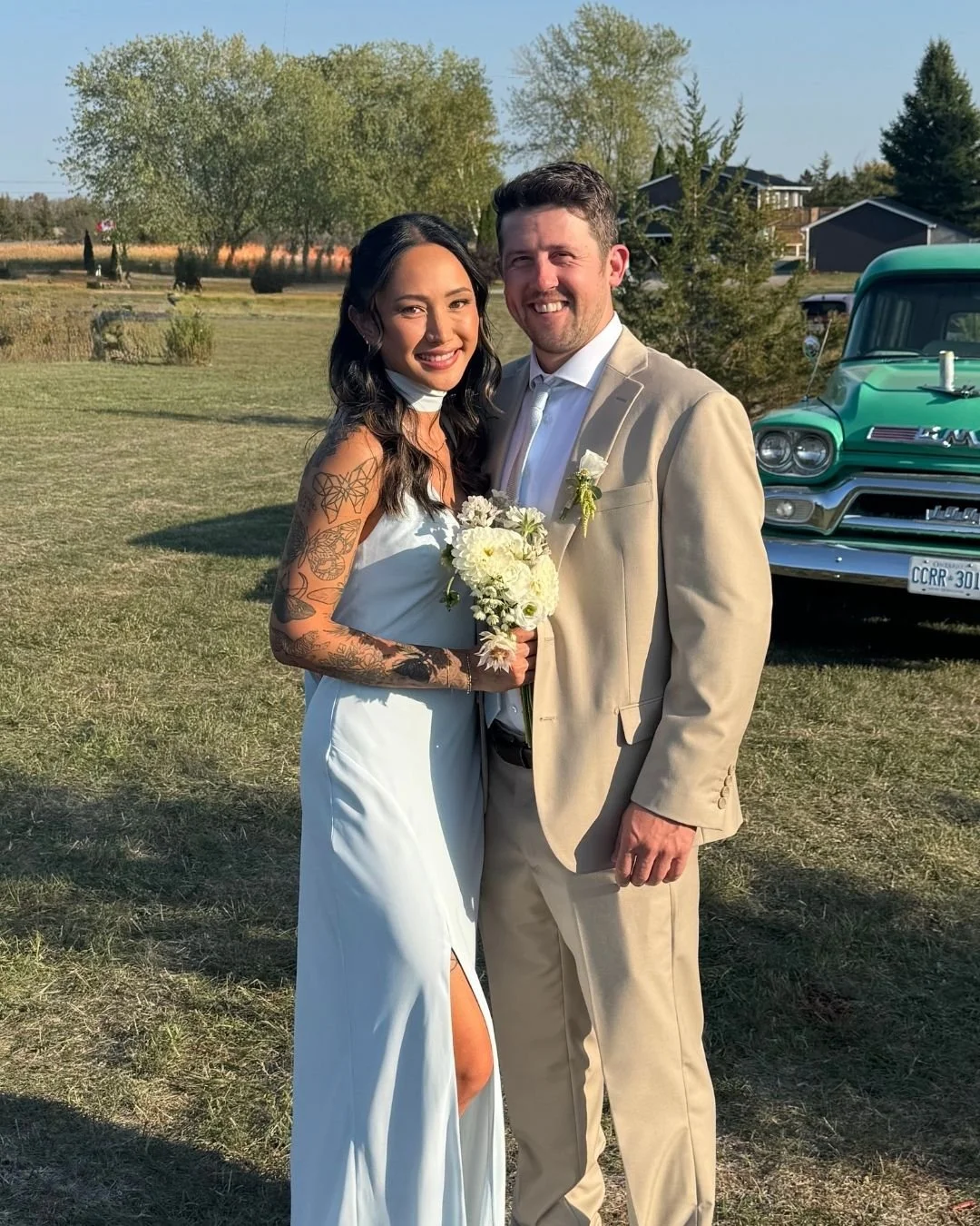 A bride and groom smiling together outdoors on their wedding day, with the bride holding a bouquet of white flowers and wearing a white wedding dress with tattoos on her arms. The groom is dressed in a beige suit, white shirt, and wearing a boutonniere. There is a vintage green truck and trees in the background.