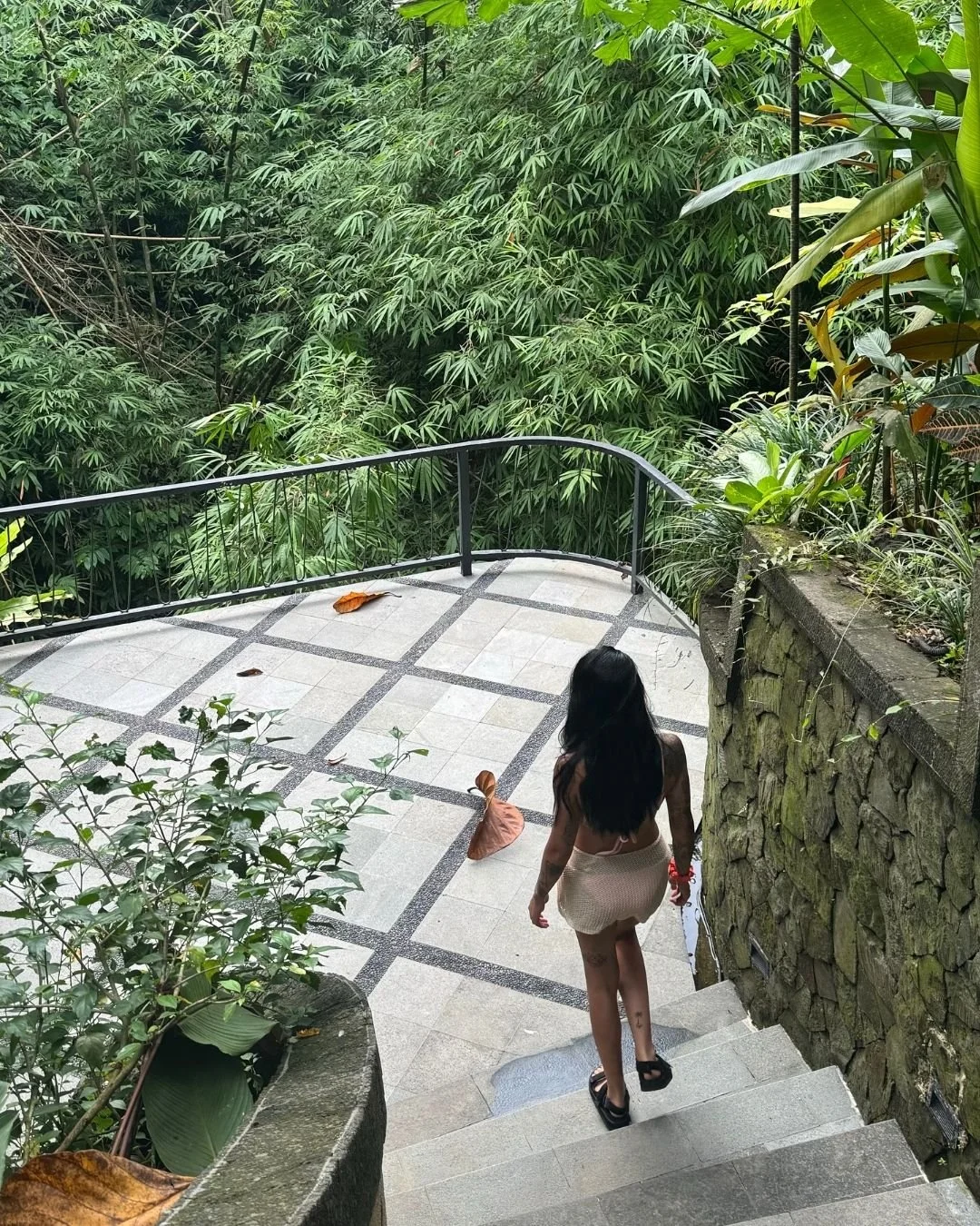 A woman with long black hair walking down outdoor stone stairs with a black railing, surrounded by lush green plants and trees, with fallen leaves on the ground.