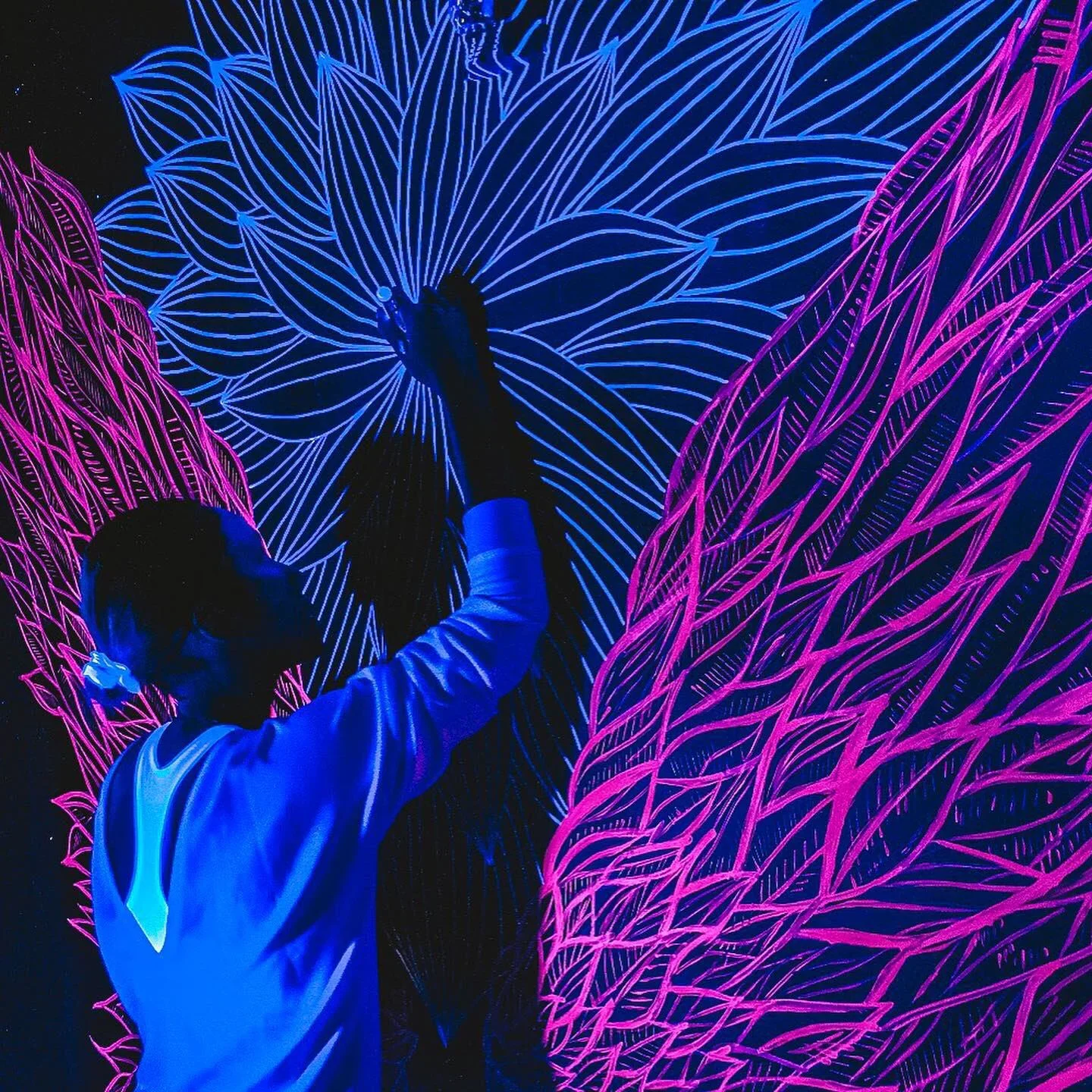 A person drawing large, detailed floral designs with neon outlines in blue and pink on a black background.