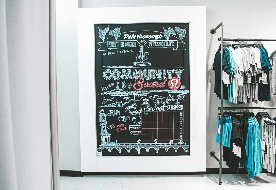 A large chalkboard sign on a white wall in a retail store. The sign has various chalk drawings and text promoting the grand opening of a community board in Peterborough, including phrases like 'What's Happening,' '#TheWeltLife,' 'Community Board,' 'S