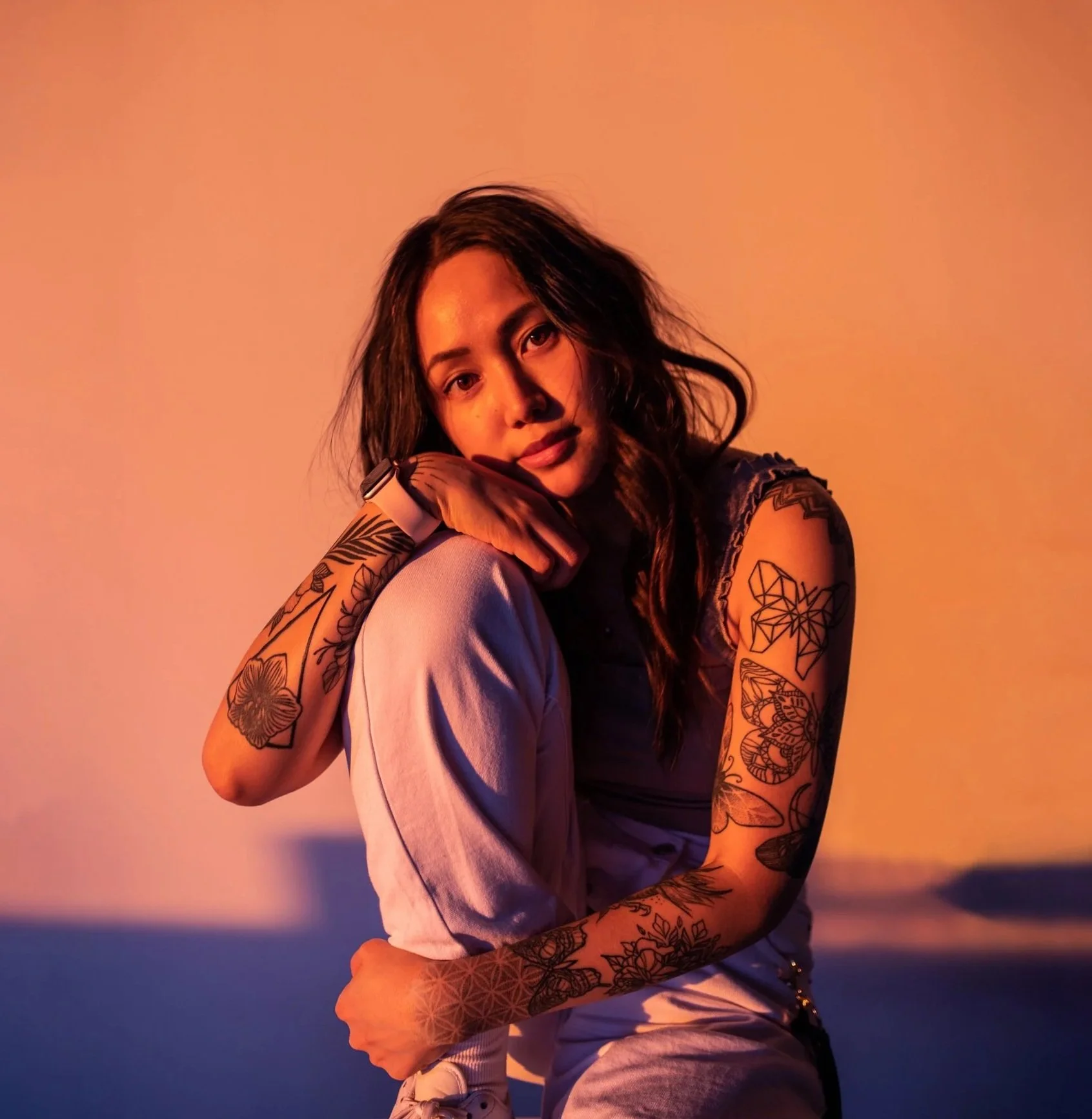 Young woman with tattoos sitting with her knee up, resting her head on her hand against a neutral background with warm and cool lighting effects.