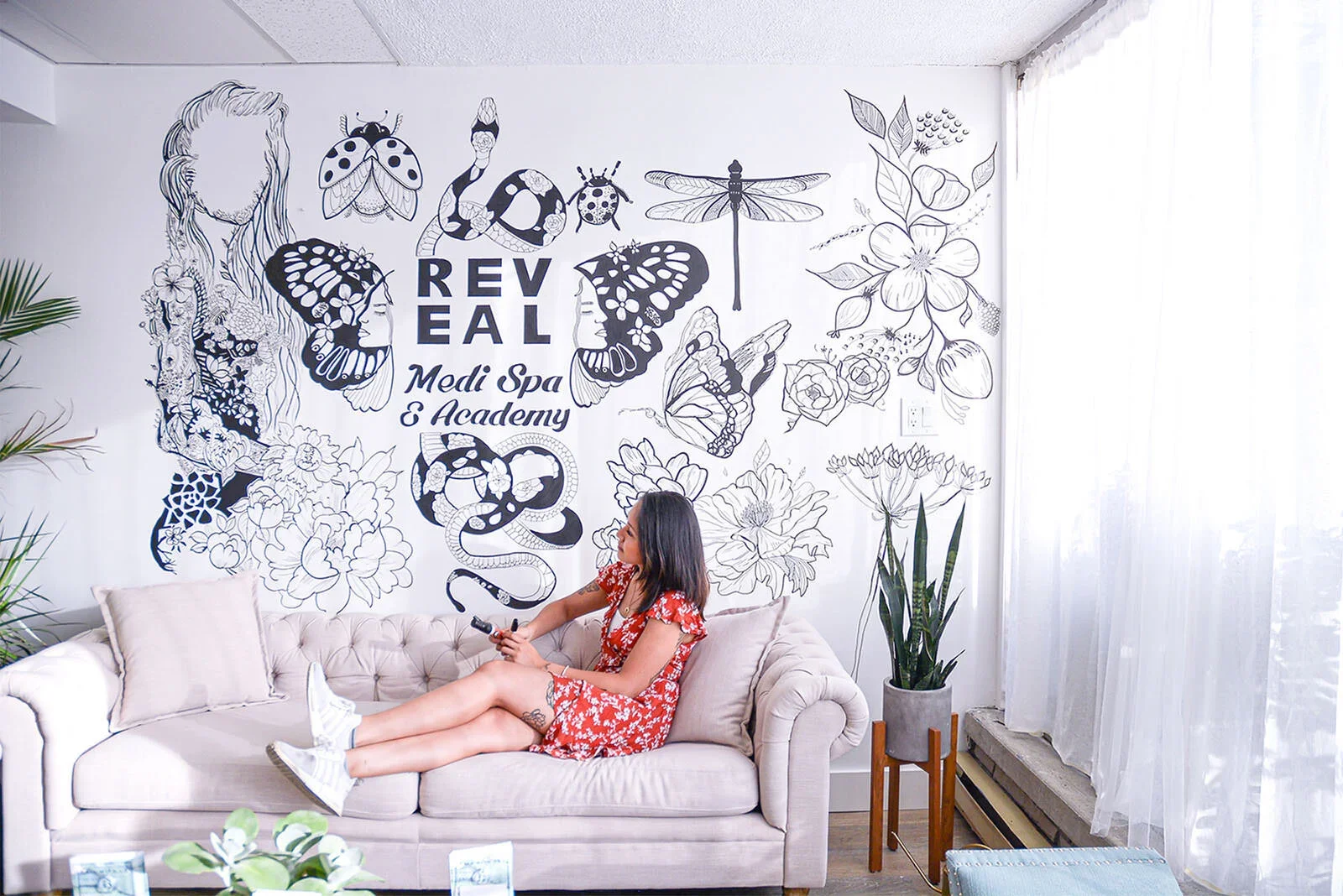 A woman in a red floral dress with white sneakers sitting on a beige tufted sofa, holding a phone, in a room with white walls and sheer curtains, styled with green plants and a wall mural featuring butterflies, insects, flowers, and the text 'REVEAL 