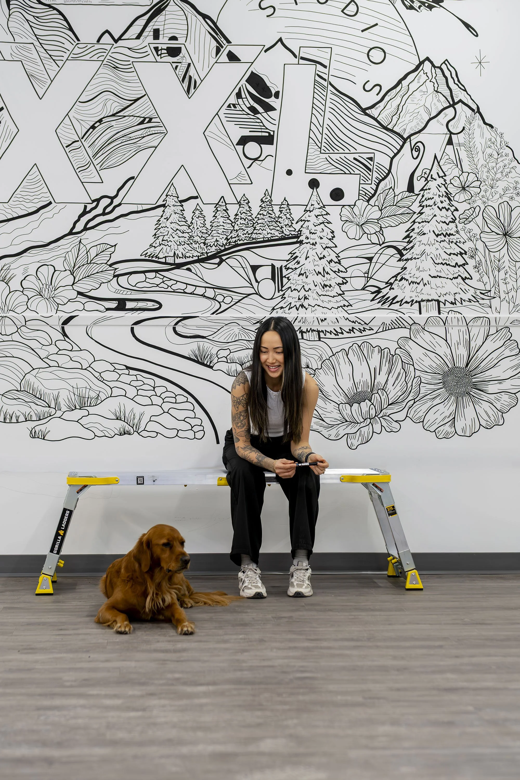 A woman with tattoos sitting on a bench with a brown dog on the floor, in front of a black and white mural of a mountain landscape with trees, flowers, a winding road, and mountain peaks.