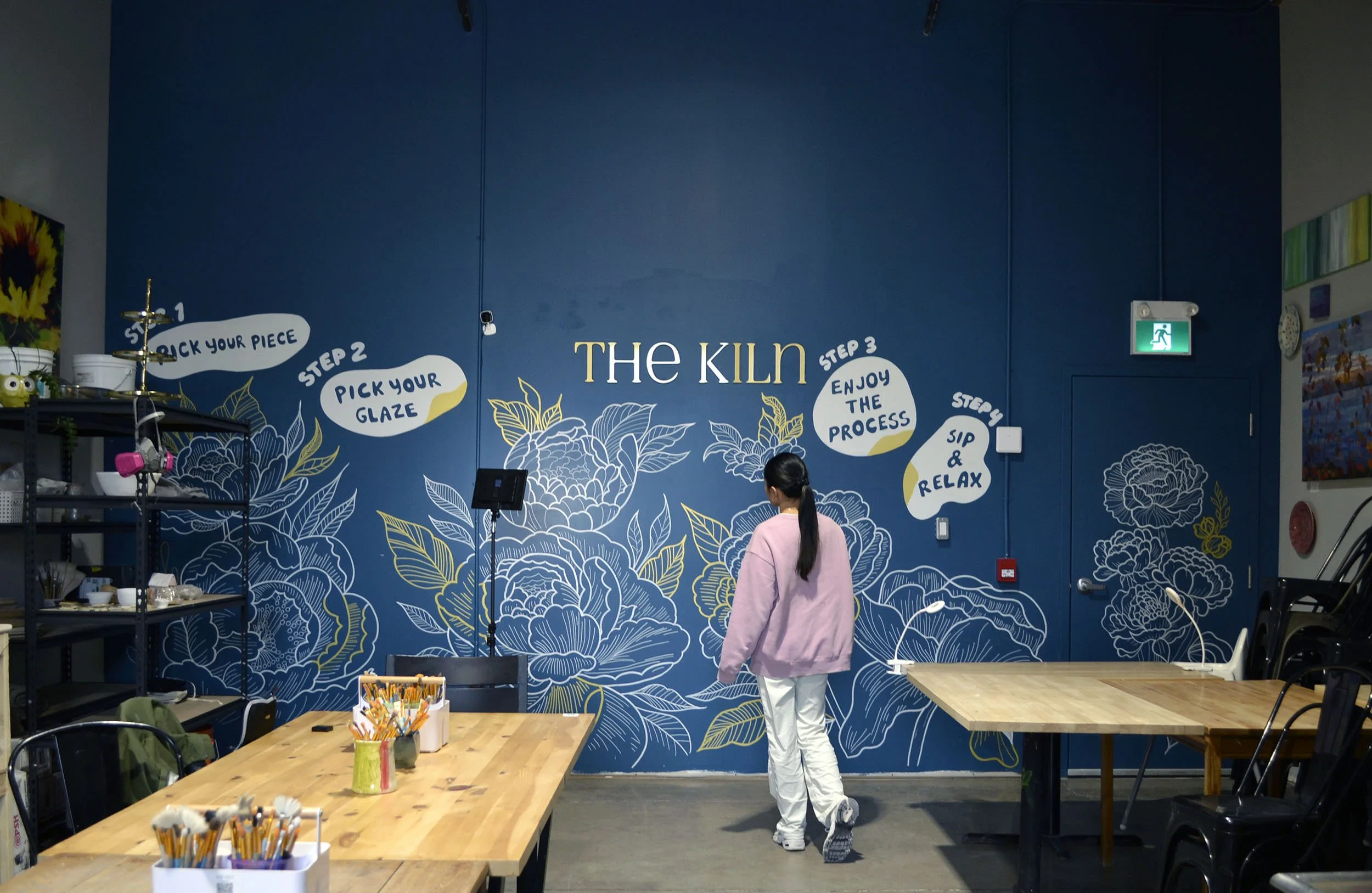 A woman in a pink sweatshirt and white pants walking in an art studio with a blue wall decorated with large floral illustrations and a step-by-step guide for a craft project, including picking up a piece, glazing, enjoying the process, and relaxing.