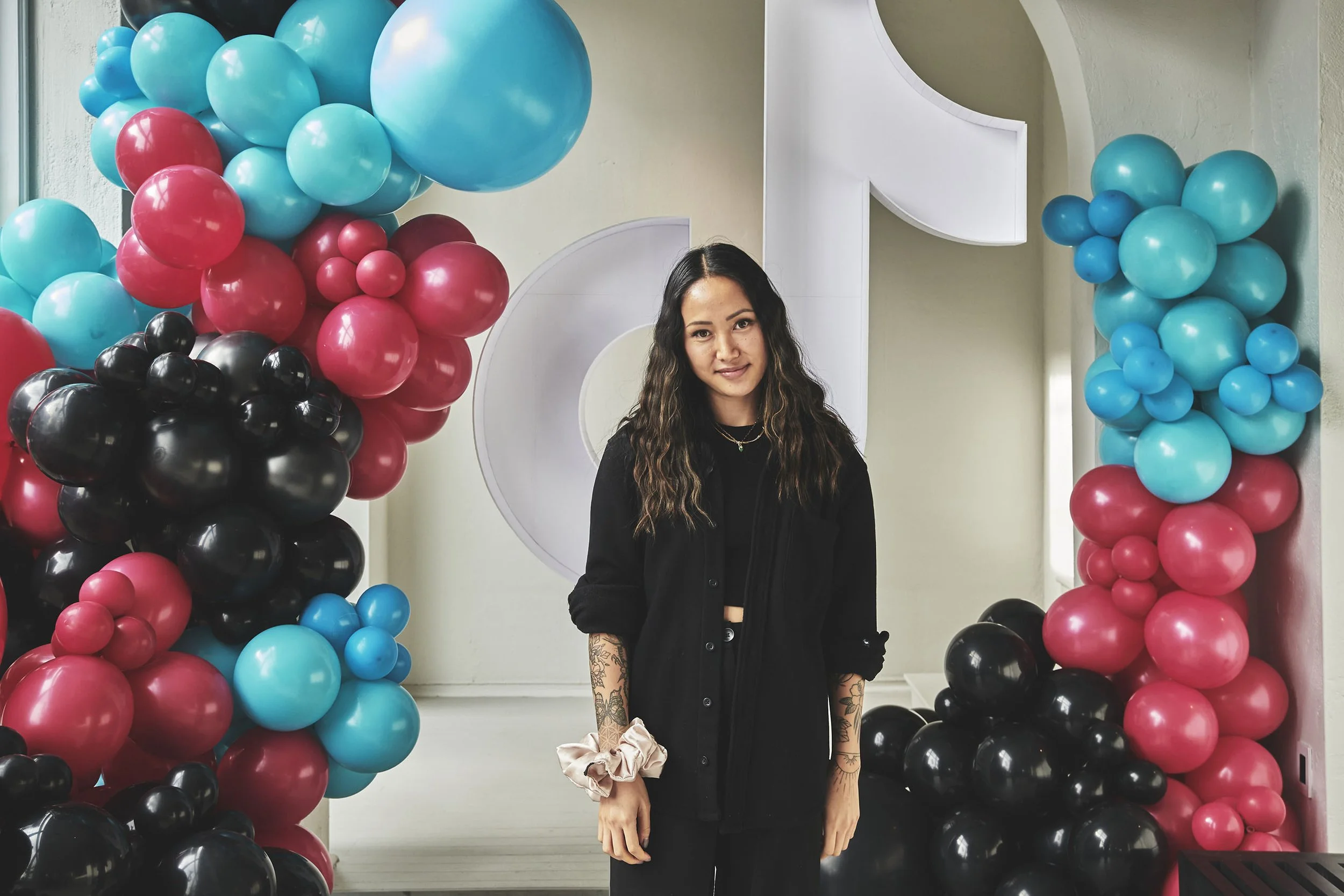 A woman with wavy dark hair and tattoos on her arms stands in front of balloon decorations in shades of blue, pink, and black.