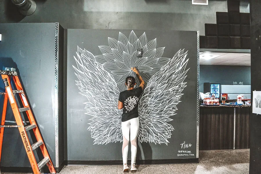 A woman drawing a large white butterfly design on a gray wall in an indoor space.