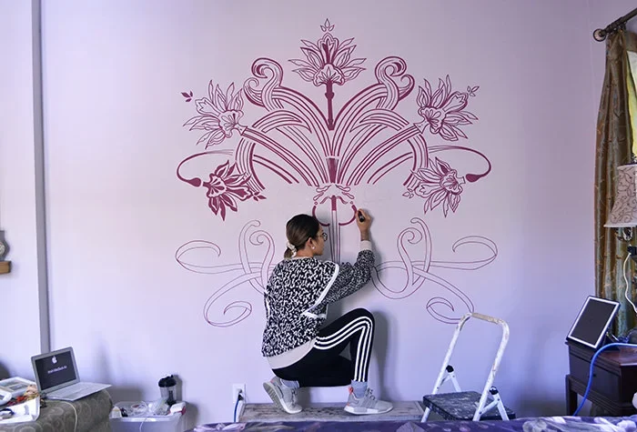 A woman drawing an intricate floral design on a white wall with a pink marker, while kneeling.