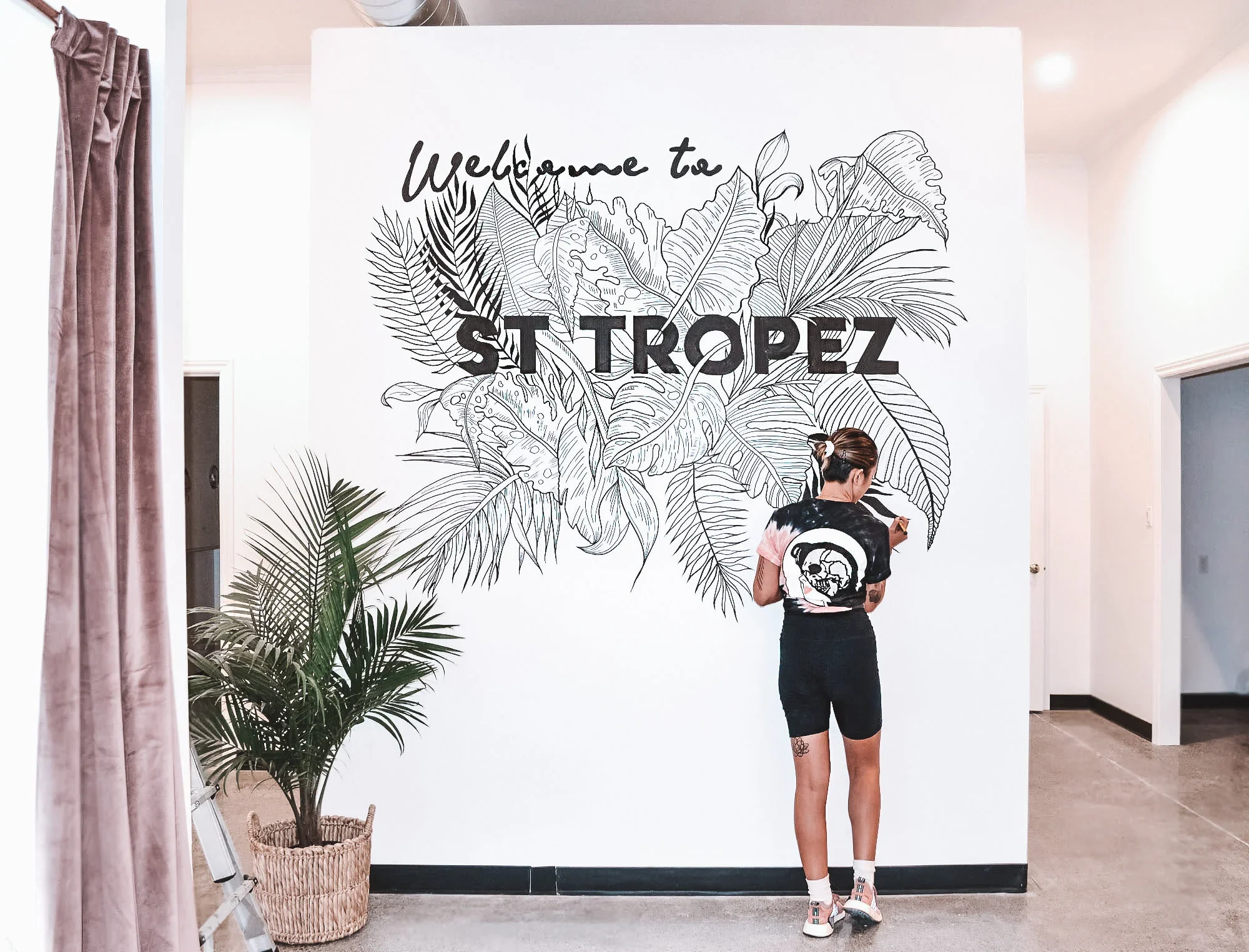 A woman standing in front of a wall mural reading 'Welcome to St. Tropez,' with large tropical leaf illustrations. She has her back to the camera, wearing a black t-shirt with a skull and rainbow design, black shorts, and sneakers. There is a potted 