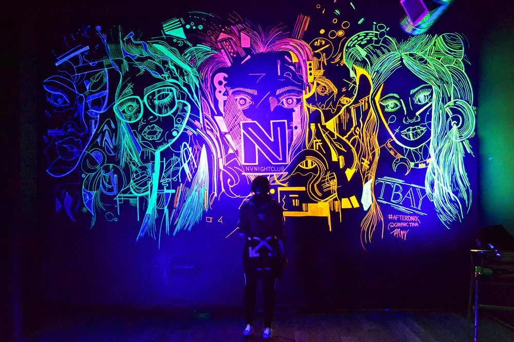 Person standing in front of a neon light wall art featuring abstract colorful portraits of women with long hair, glasses, and accessories at Itty Bitty in New York City.
